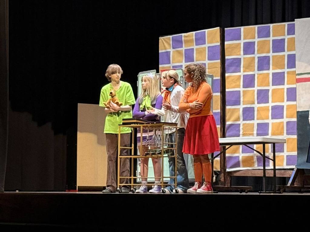 Four individuals on a stage, two holding items, one in a red skirt, and a table and chairs.