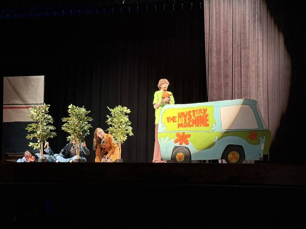 On a stage, performers act out a scene. A person stands on a vehicle with text. Potted plants are nearby.