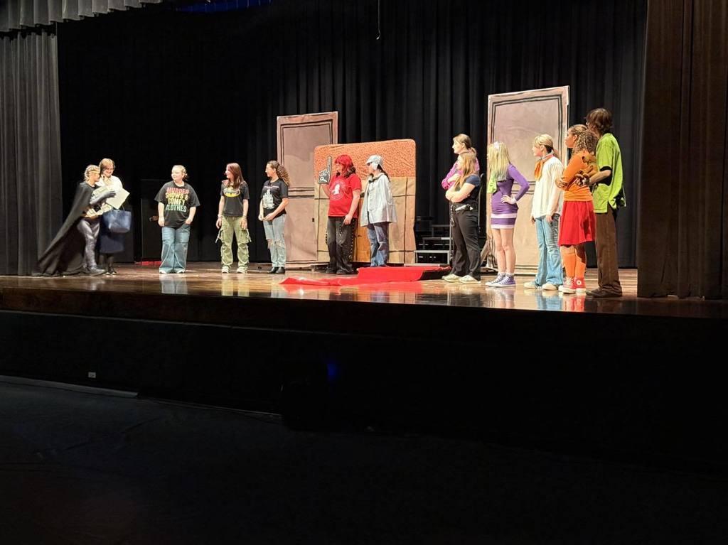 A group of people stands on a stage, illuminated by a spotlight. Some wear costumes; two large doors are in the background.