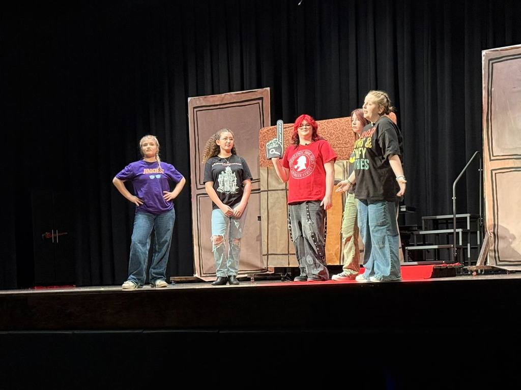 Five people on stage facing the audience. One person holds a microphone. Background includes doors and black curtains.