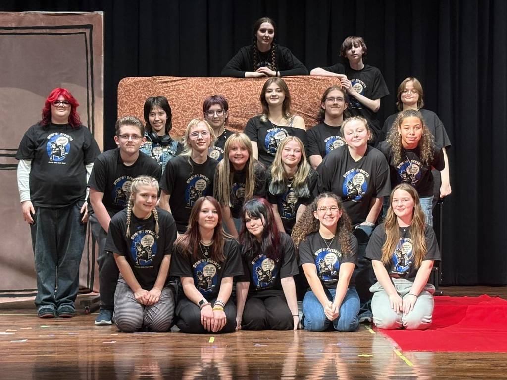 A group of individuals in matching black t-shirts are on a stage, with some sitting and others standing behind a large brown prop.