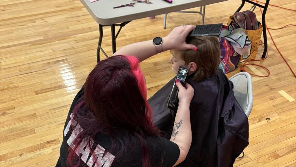 A woman trims a young child's hair in a school gym. The child sits in a chair.