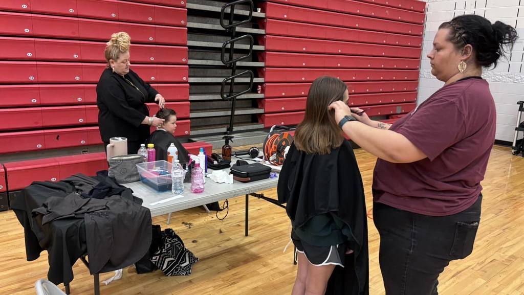 Three women in a gymnasium with red bleachers. One woman sits in a chair getting her hair cut.