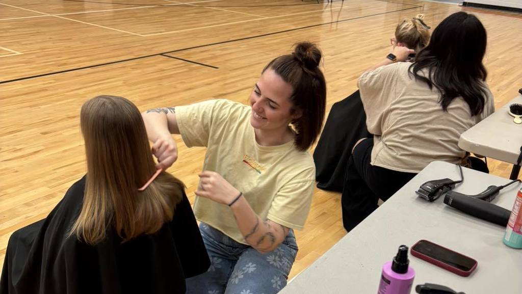 A person cuts another's hair in a gymnasium. Other people sit at tables. A basketball court is in the background.