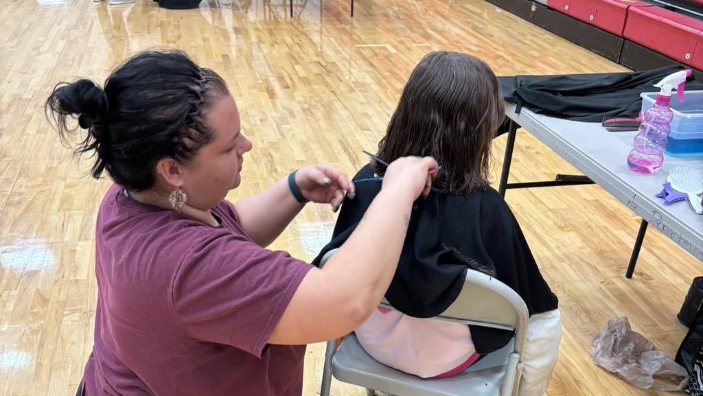 A woman is cutting a young girl's hair in a chair. A table with bottles is nearby.