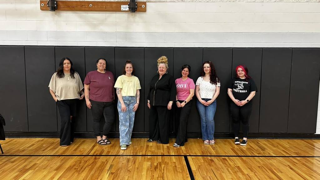 Seven people in different tops and pants, likely a team, stand on a basketball court with a black wall.