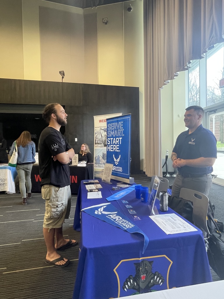Two men are at a table with a blue cloth, talking. A blue banner is behind them. People walk past them.