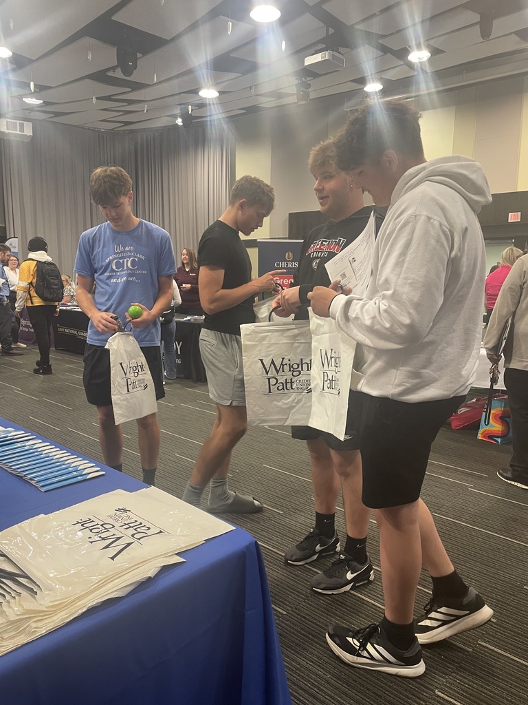 Four people stand at a table with bags. One person holds a tennis ball. A blue table holds printed material.