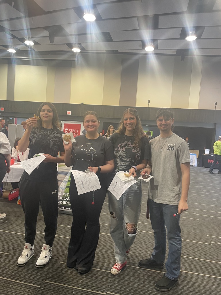 Four people stand together in a room, holding certificates. They are wearing sneakers and casual clothes. A blurred background shows a person in the distance.