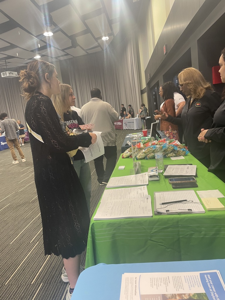 People in a large room are gathered around a green table with items on it and papers.