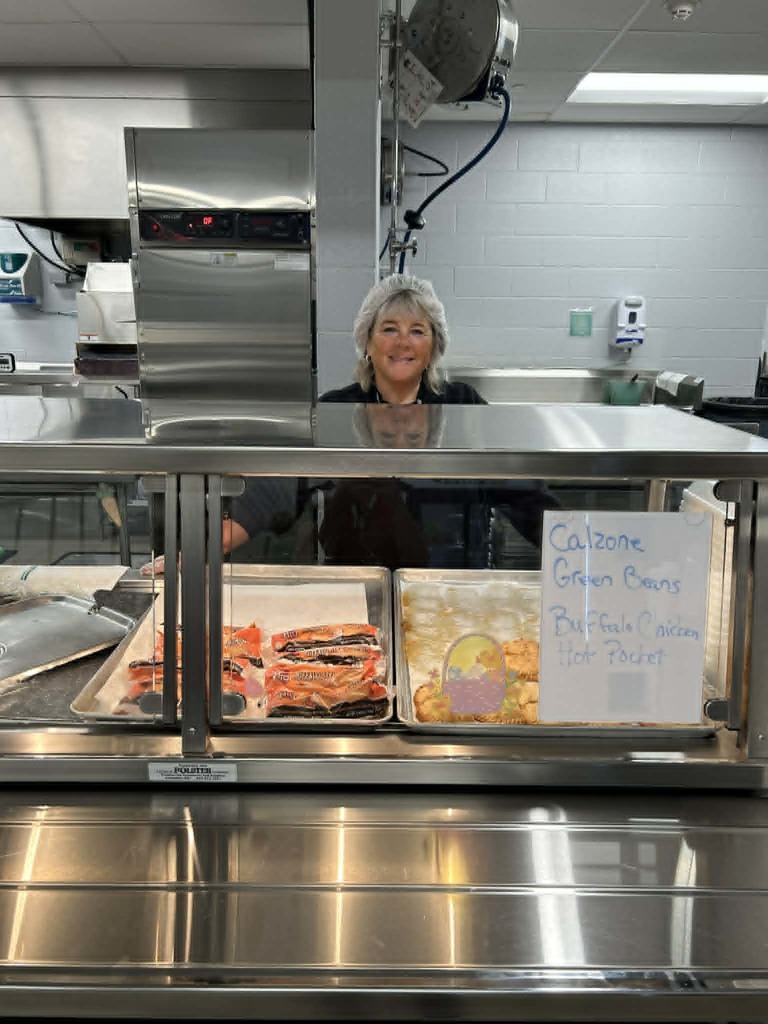 A woman stands behind a counter in a kitchen with trays of food. A sign reads "Calzone Green Beans Buffalo Chicken Hot Pastrami."