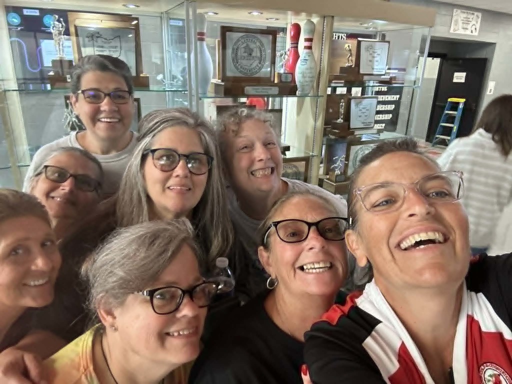 A group of people wearing glasses and smiling, standing in a bowling alley with bowling trophies in display cases.
