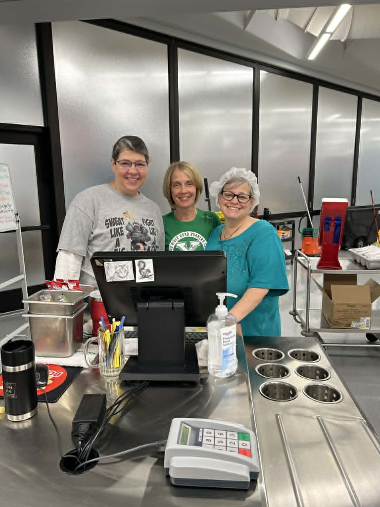 Three women stand behind a counter, with a computer and various items on the counter, including a mug and a bottle.