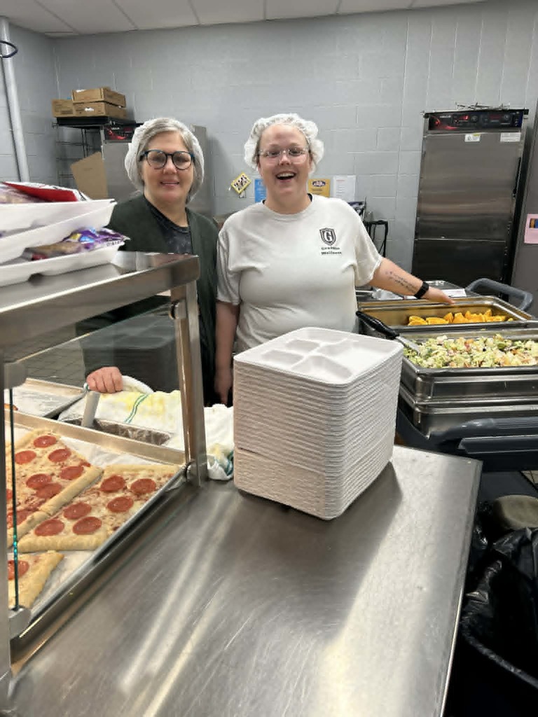 Two women in a kitchen wearing hair nets stand behind a table with pizza, plates, and trash.