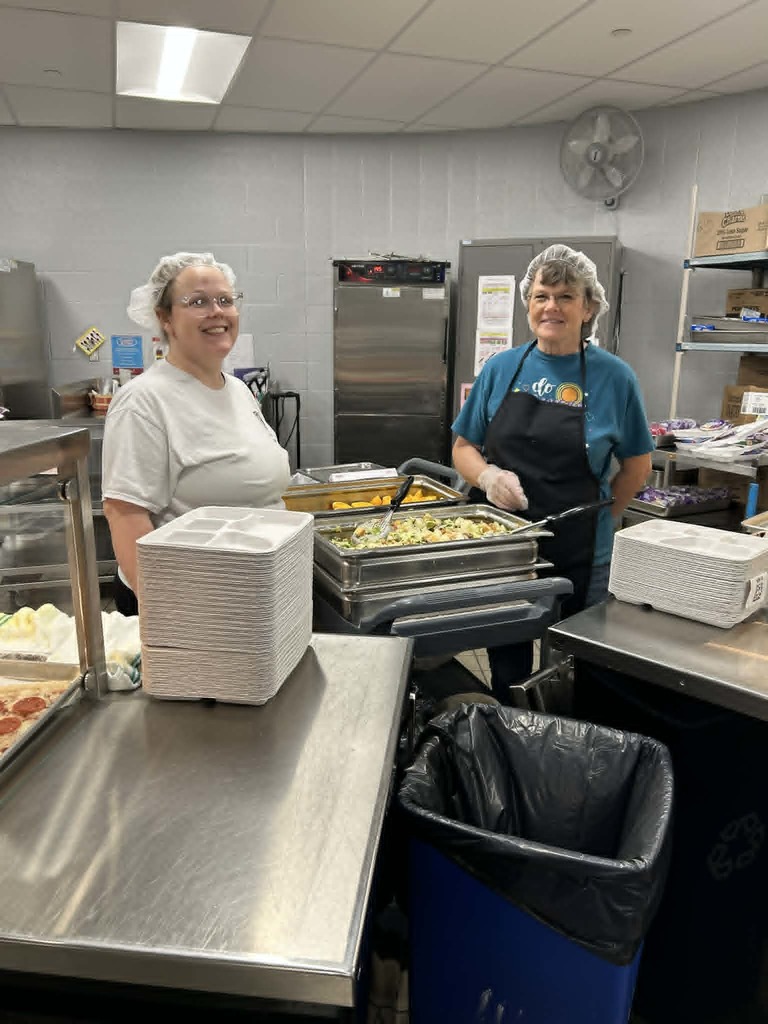 Two women in a commercial kitchen, one wearing a blue apron, standing near trays and a trash bin.
