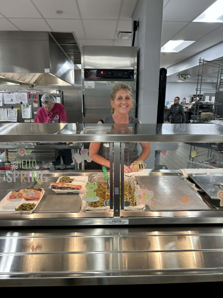 Woman stands behind a display case of food in a commercial kitchen with other workers.