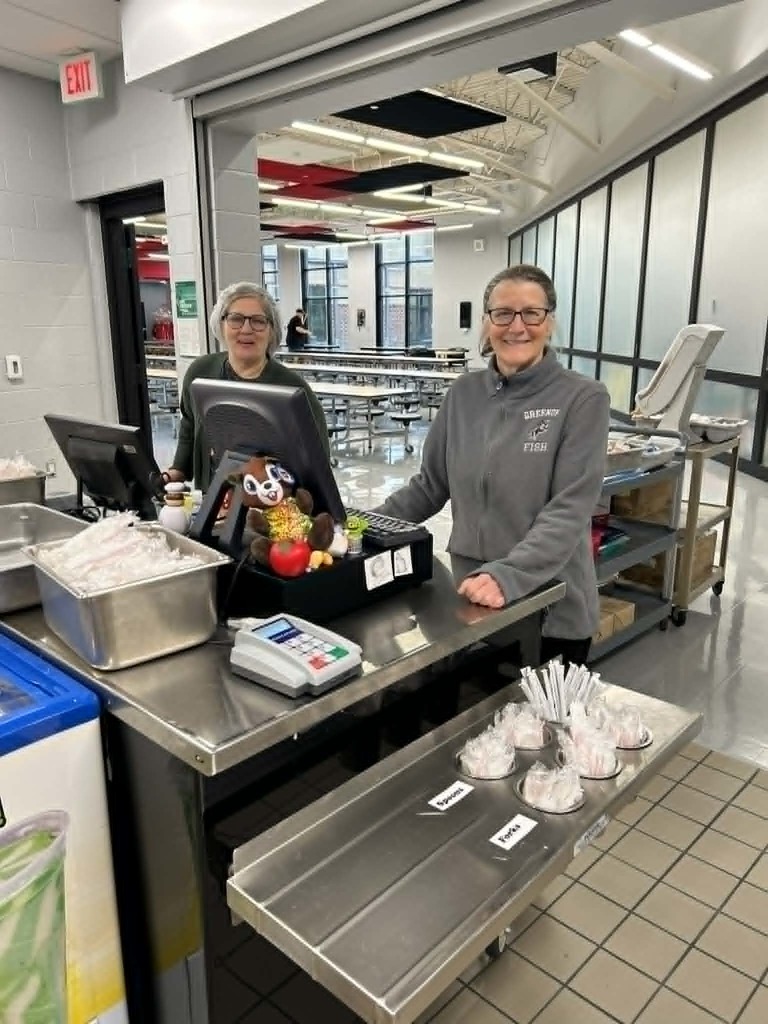 Two women stand behind a counter with food items and utensils. A computer is on the counter.