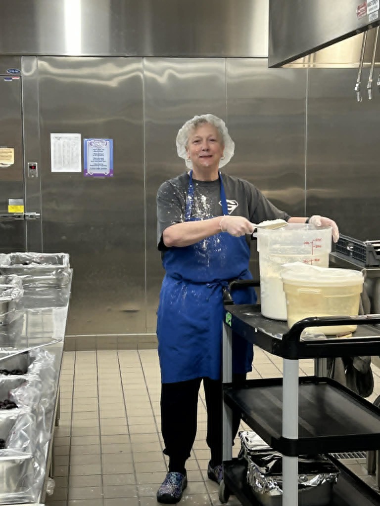 Person in blue apron standing in a commercial kitchen, holding a plastic container. Stainless steel walls and tiled floor.