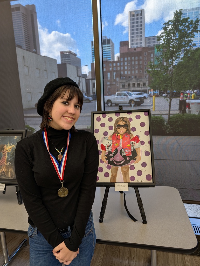A woman stands beside a painting on an easel. She wears a medal and stands in a room with large windows showing a city.