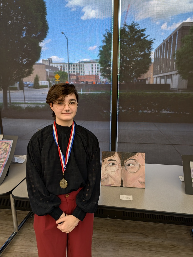 Person wearing a medal stands in front of a display table with artwork. They stand in front of a window.