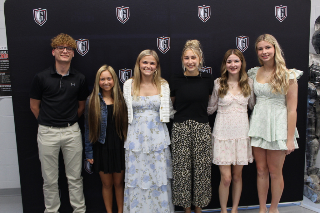 Six young people in various dresses and one in a black shirt and khaki pants stand in front of a wall.