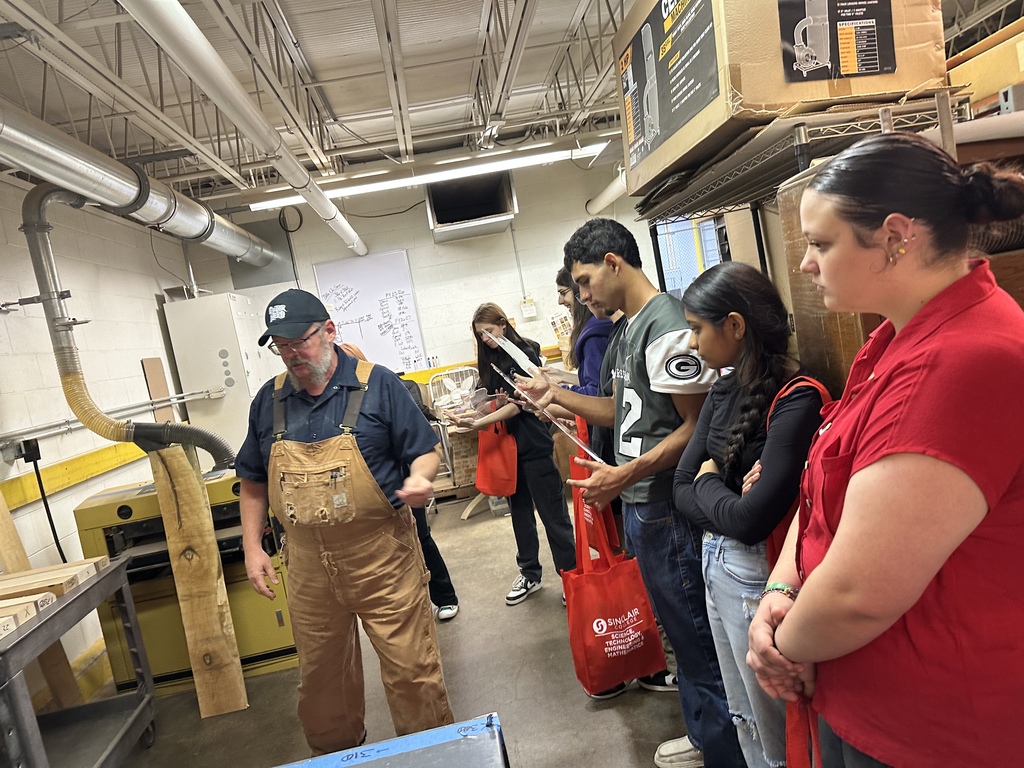 A man in overalls talks to a group of students, some holding bags, in a factory.