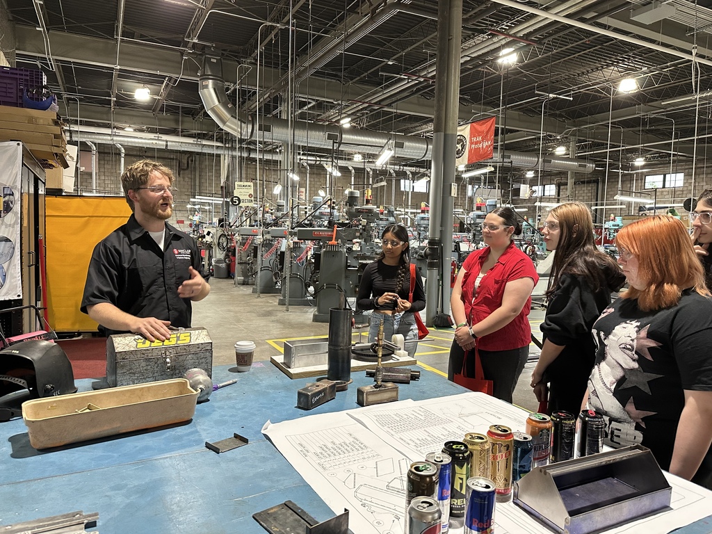 Factory with tall ceilings. A man in a black shirt speaks to a group of people.
