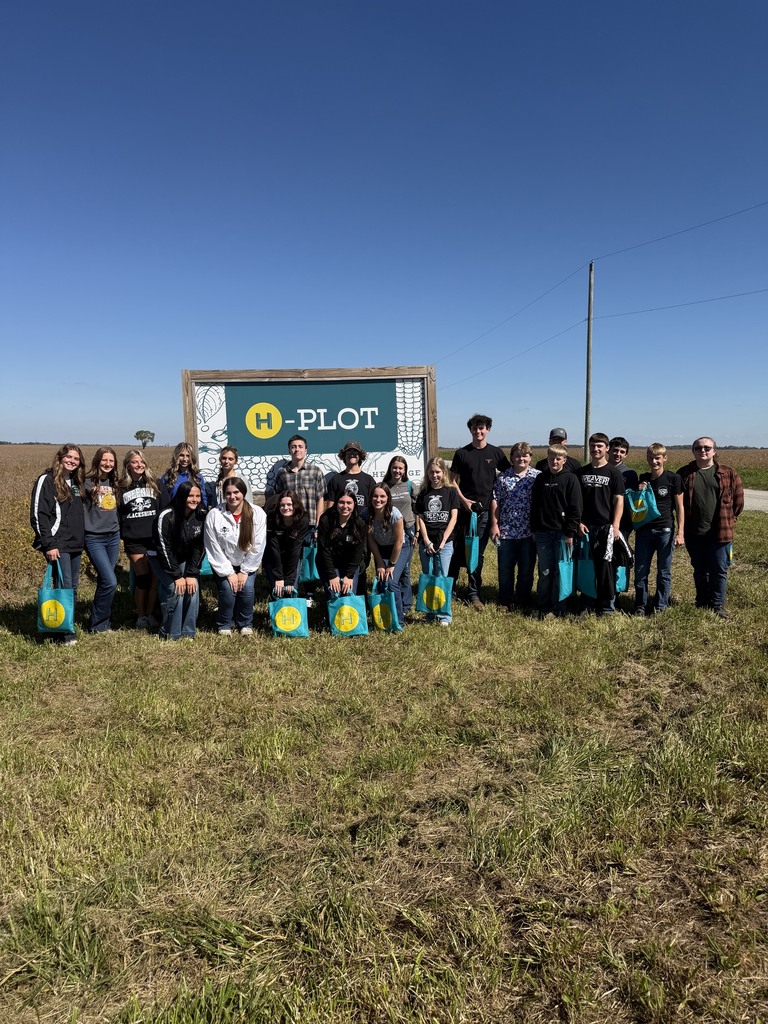 Group of people posing for a photo in a field with tall grass and a sign reading "H-PLOT".