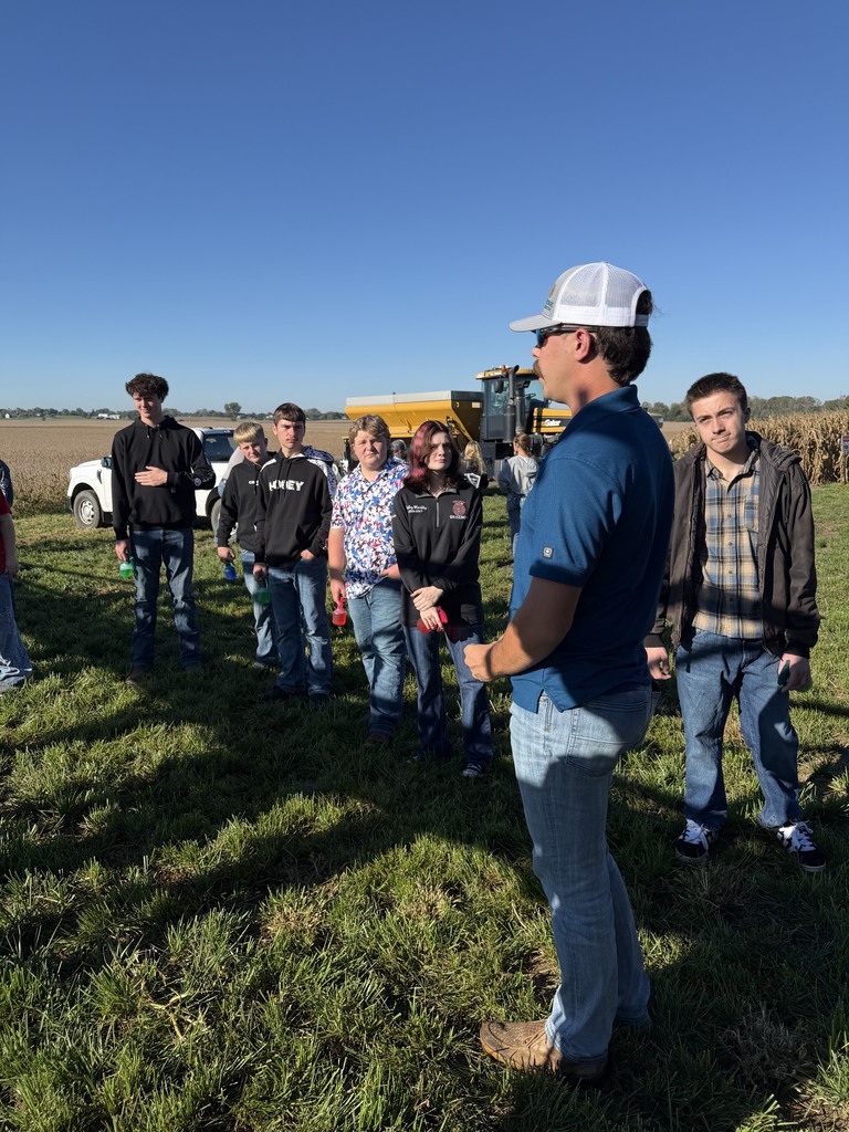 A man wearing a cap speaks to a group of people in a grassy field. They are near a vehicle.