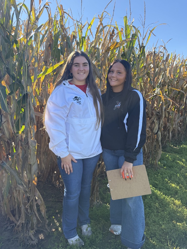 Two women stand in a cornfield, one holding a clipboard, with tall corn stalks behind them and a clear blue sky.