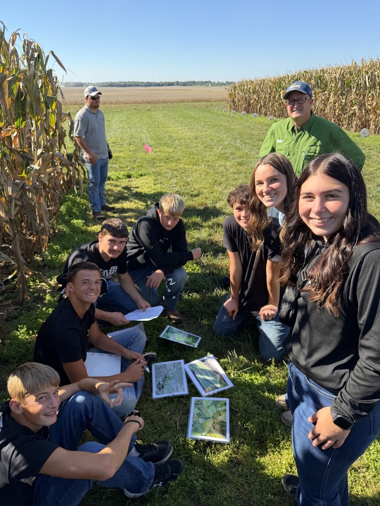 People sit in a field. Some hold papers and smile. Corn grows tall in the background.