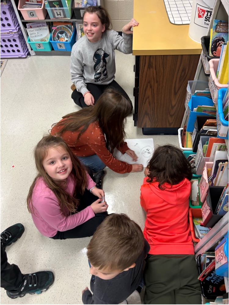 5 students working together on a sketching a snowman on white paper with pencil, one student is looking at the camera smiling