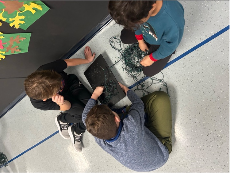 3 males students work on stringing lights into a small black billboard to recreate their snowman sketches
