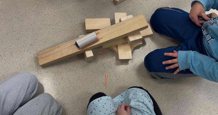 students are out of frame, but their hands are shown testing their bobsled on a built wooden ramp
