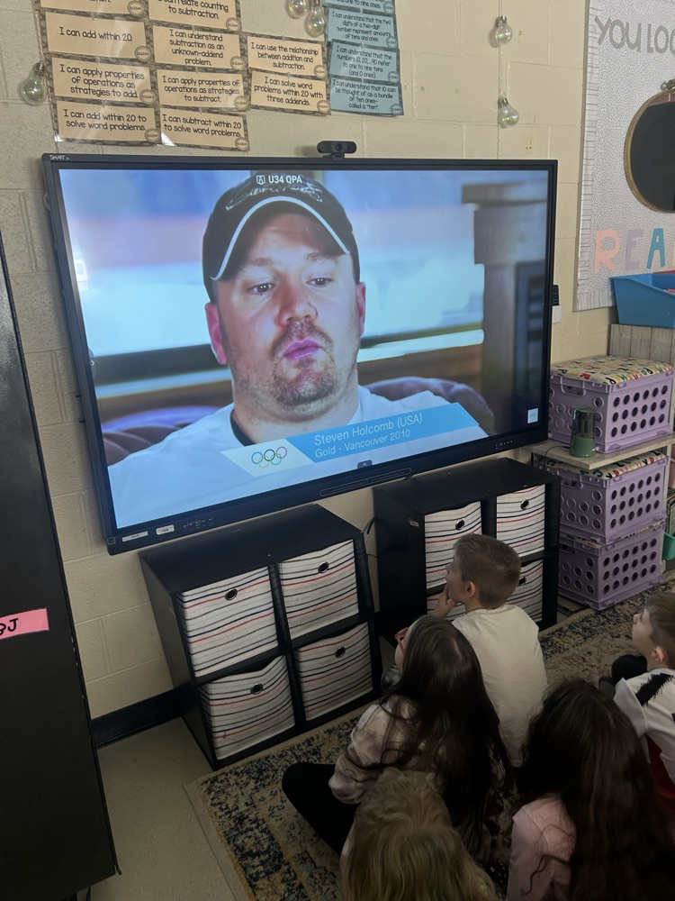 students sit on a carpet in front of their smart-board watching a video of a bobsleigh Olympian  