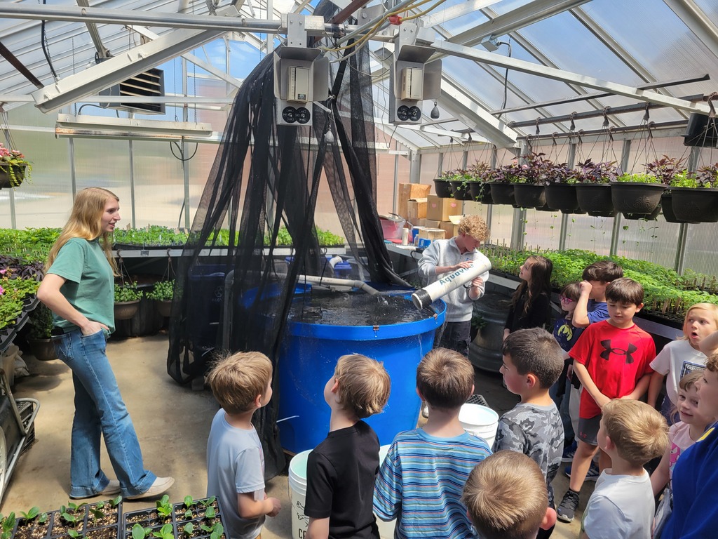 Inside a greenhouse, children stand around a large blue water tank while one person holds a view finder into the tank, with hanging planters, seedling trays, and greenhouse equipment visible overhead.