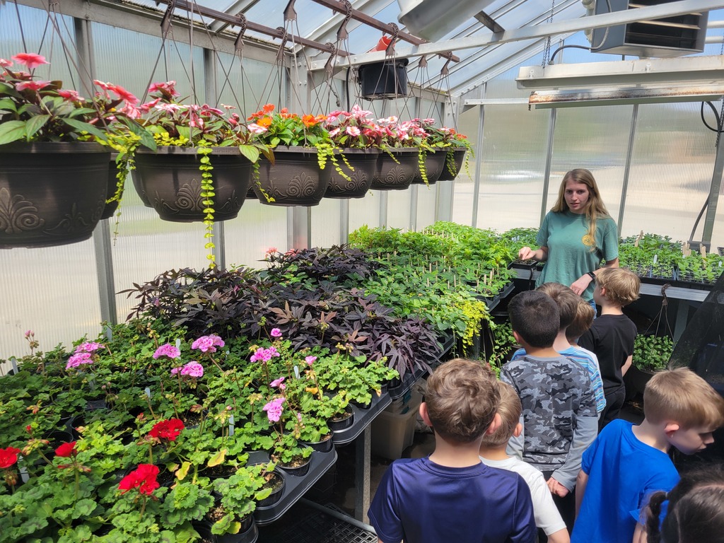 Inside a greenhouse, rows of green plants and flowering seedlings sit on tables beneath hanging baskets filled with pink and orange flowers, while a high school students stands talking with a small group of children gathered along the aisle.