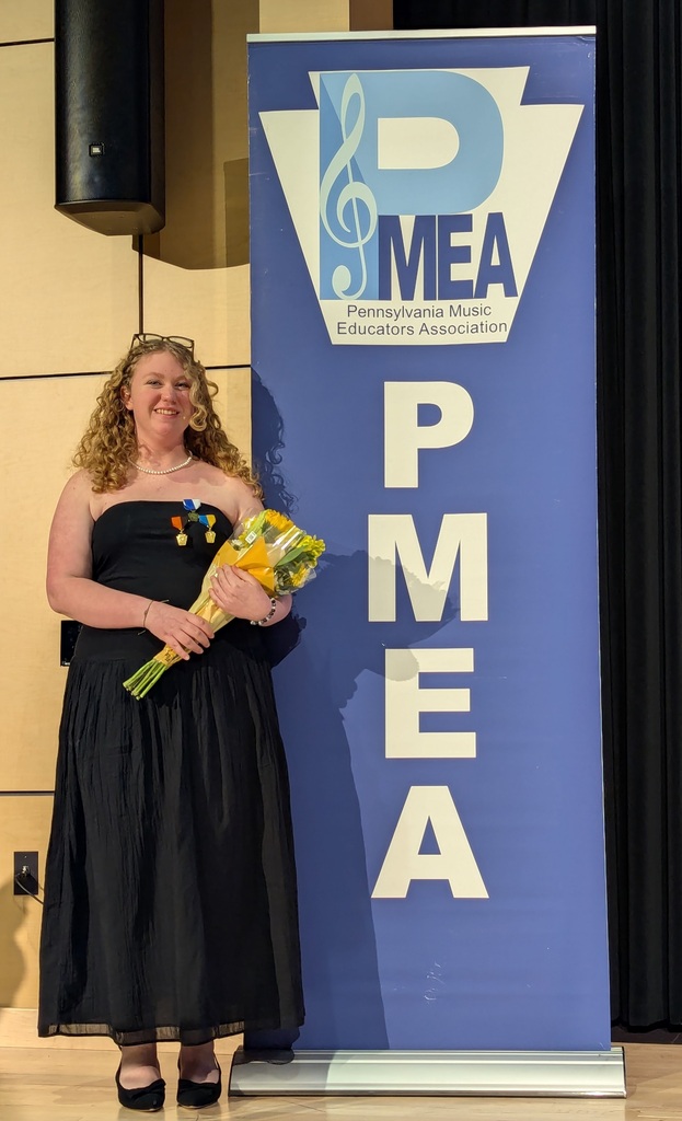 Student holding flowers and smiling next to PMEA banner at music festival.