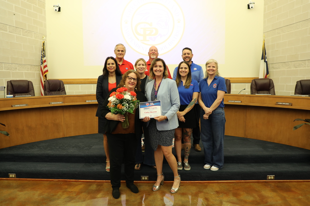 Group of people standing in a school board meeting room, presenting a certificate and bouquet for a community recognition in front of a podium and flags.