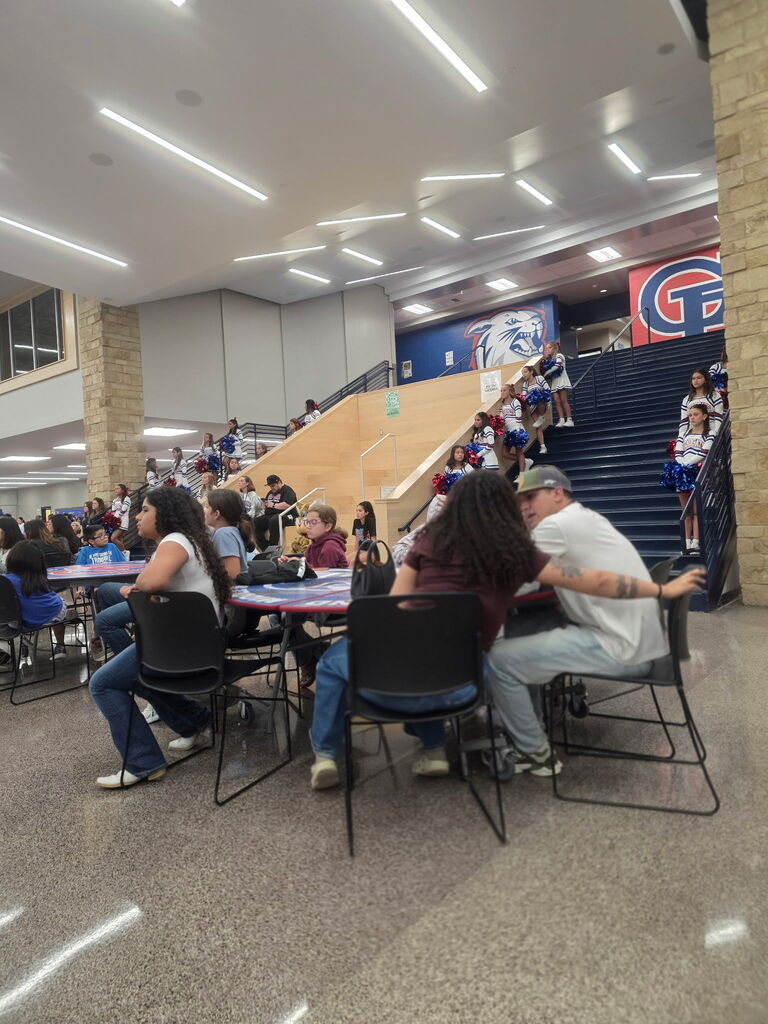 A crowded cafeteria-style seating area with people sitting at tables and benches, some eating and others conversing.