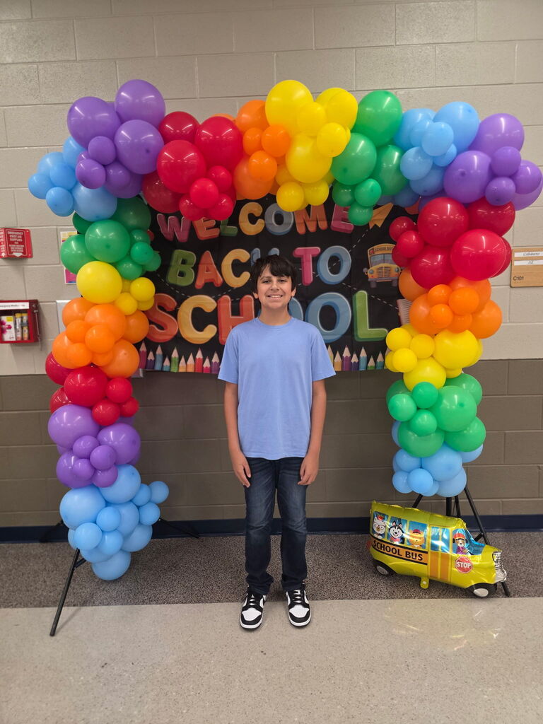 Colorful balloon arch with "Welcome Back to School" text, and a smiling person standing in front of it, holding a school bus toy.