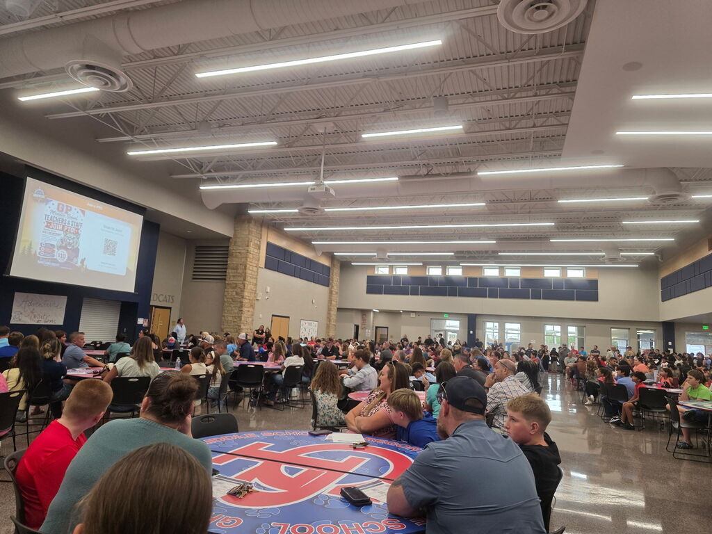 A crowded indoor event or activity with many people seated at tables and looking at a screen at the front of the room.
