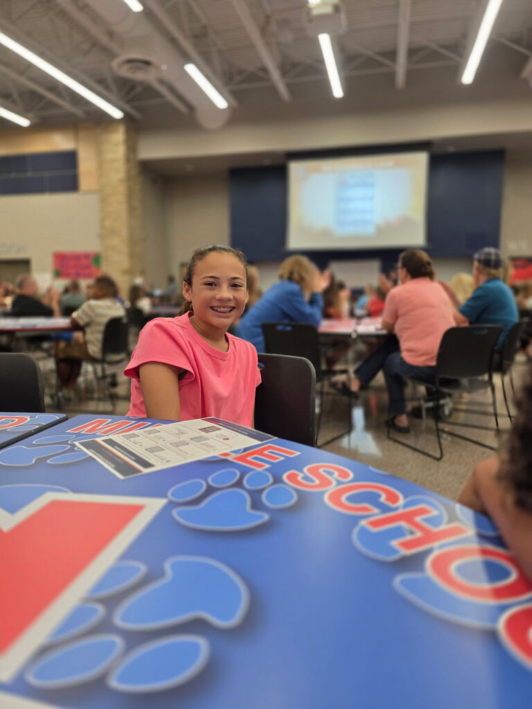 Smiling girl seated at a blue-painted table in a classroom setting, with rows of students and a projection screen visible in the background.
