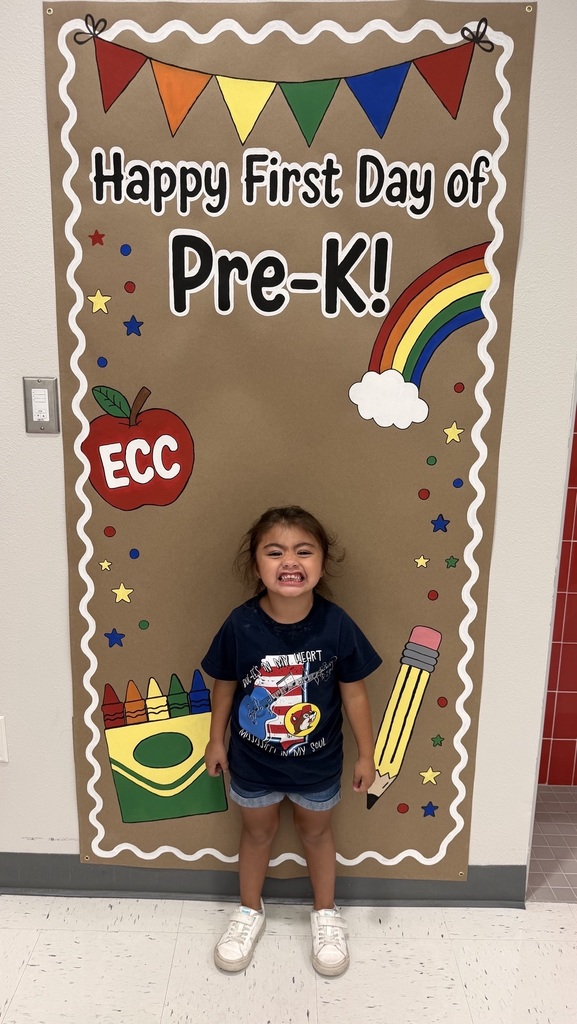 A young child smiling in front of a colorful "Happy First Day of Pre-K!" sign with an apple, crayons, and a rainbow.