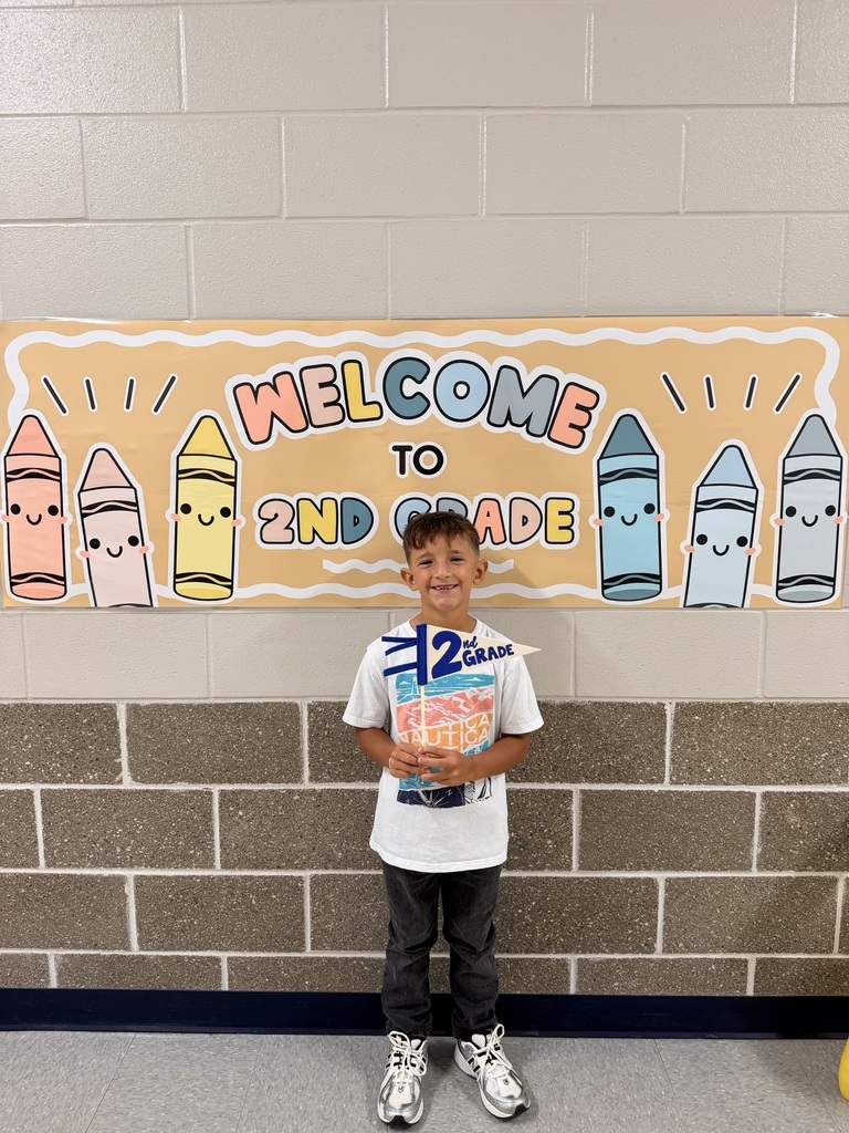 A young child standing in front of a "Welcome to 2nd Grade" banner with decorative pencils.