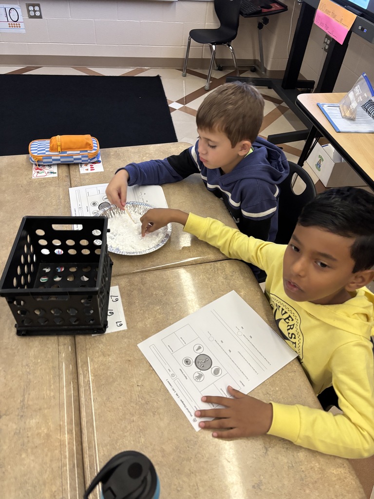 Two young students in a classroom working on an assignment together, one holding a worksheet and the other manipulating objects on the desk.