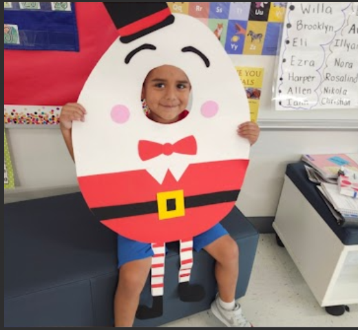 A smiling child wearing a Humpty Dumpty costume in a classroom setting, with a whiteboard and other student artwork visible in the background.