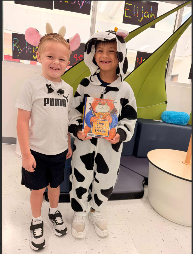 Two smiling children in animal-themed costumes, one wearing a cow onesie and holding a book, the other in a plain t-shirt.