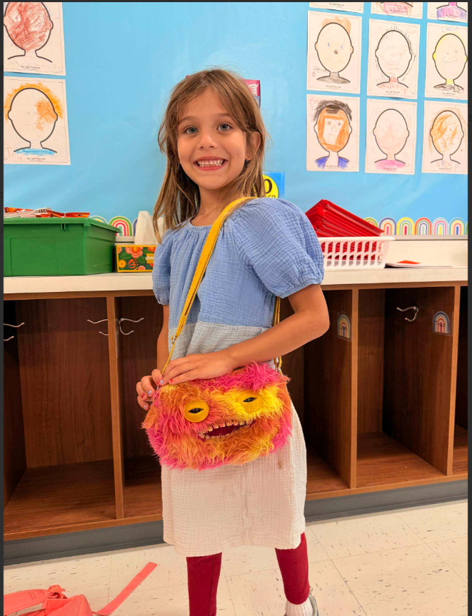 A young girl with blond hair wearing a blue shirt and holding a colorful, fuzzy purse in a classroom setting with shelves and artwork.