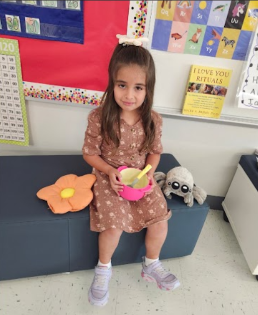 Young girl in floral dress sitting on couch in classroom, holding plush toys and smiling. Bulletin board with visuals and text displayed on wall behind her.